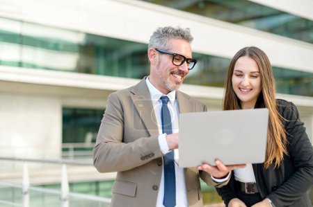 A mature businessman and his female colleague looking at a laptop screen standing outdoors together, with a clean architectural background emphasizing the fusion of business and modern design.