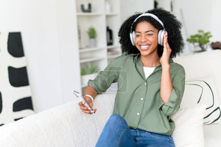 Eine fröhliche Afroamerikanerin mit Kopfhörern sitzt gemütlich auf einem Sofa und hält ein Smartphone in der Hand. Das Konzept fängt einen entspannten, fröhlichen Moment des Genießens von Musik oder einem Podcast zu Hause ein.