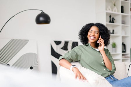 The woman relaxes on the couch, her contagious smile present as she enjoys a casual conversation over the phone, epitomizing the comfort and connection of mobile communication