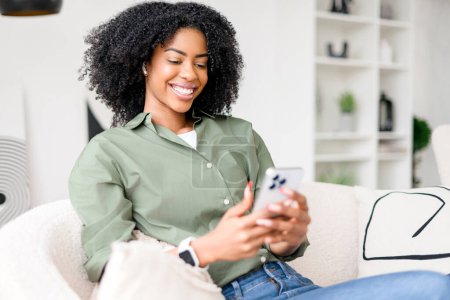 Una joven afroamericana emana felicidad mientras usa su teléfono inteligente, compartiendo una risa en una conversación digital, mientras se encuentra en una sala de estar elegantemente decorada
