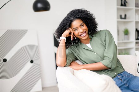 Mit einer Hand auf der Wange zeigt eine afroamerikanische Frau einen entspannten und nachdenklichen Ausdruck, während sie in einem modernen Wohnzimmer sitzt und einen ruhigen Moment häuslicher Glückseligkeit widerspiegelt..