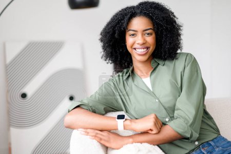 Assise de façon décontractée sur un canapé, une femme afro-américaine porte un sourire délicieux, les bras croisés confortablement, reflétant une aura de simplicité et de vie moderne dans une chambre décorée avec goût..