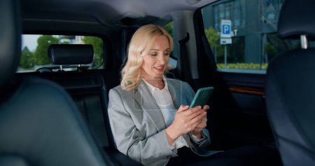 Photo for Good-looking caucasian woman in formal clothes with smartphone on the back seat of a car. Middle-age woman uses a mobile phone in the car. Internet and social media. Lifestyle - Royalty Free Image