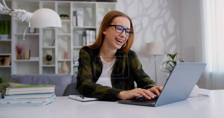 Attraktiv lächelnde moderne blonde junge Frau, die zu Hause am Schreibtisch sitzt und mit Freunden am Computer plaudert, Freizeitkonzept