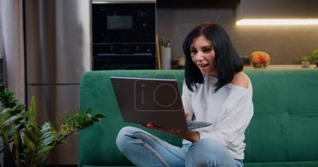 Woman reading good news on laptop screen at home. Winner amazed female sits in kitchen celebrates victory wins online sales discounts internet auction bid success.