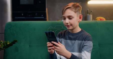 Portrait of a little boy using smartphone watching to screen, reading, typing, playing games sitting on sofa at home.