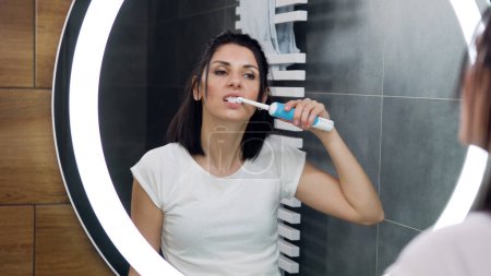 Téléchargez les photos : Attrayant concentré jeune femme en vêtements domestiques regardant dans le miroir rond tout en se nettoyant les dents avec une brosse à dents - en image libre de droit
