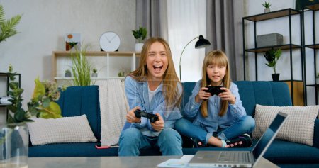 Téléchargez les photos : Gros plan de assez satisfait excité deux filles aînées et jeunes sœurs qui sont assis sur un canapé confortable à la maison et profiter de jeux vidéo appliquant joysticks - en image libre de droit
