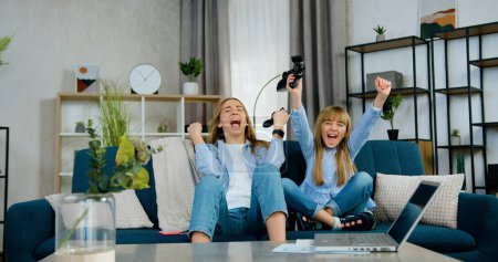 Téléchargez les photos : Belle émotionnelle positive deux âges différents sœurs célébrant la victoire dans les jeux vidéo avec les mains levées, puis donner cinq élevé l'un l'autre - en image libre de droit