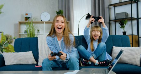Téléchargez les photos : Belle amicale gaie deux âges différents filles jouer à des jeux vidéo en utilisant des joysticks et célébrer la victoire avec les mains levées lors de la dépense conjointe de temps libre à la maison - en image libre de droit