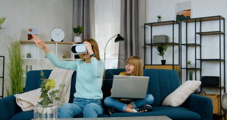 Photo for Beautiful positive modern two different ages girls spending joint leisure at playing virtual games using laptop and augmented reality goggles,front view - Royalty Free Image