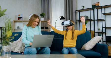 Photo for Charming joyful light haired 20-aged girl choosing virtual entertaintment for her younger sister which sitting near her on soft couch in augmented reality goggles - Royalty Free Image