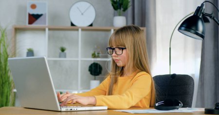 Téléchargez les photos : Vue latérale d'agréable confiant concentré fille de 10 ans dans des lunettes qui sont assis devant l'ordinateur et honnêtement faire ses devoirs - en image libre de droit