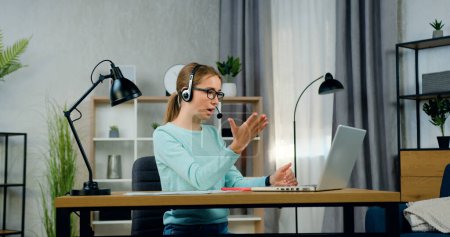 Charmantes, intelligentes 20-jähriges hellhaariges Mädchen mit Kopfhörern, das am Tisch im modernen Wohnzimmer sitzt und per Videochat auf dem Laptop mit seinen Gesprächspartnern spricht