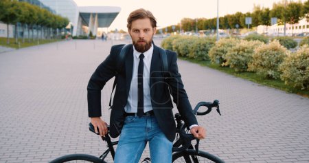 Foto de Primer plano de guapo hombre moderno y elegante con barba que se sienta en su bicicleta en el camino peatonal cerca de la construcción urbana grande y mirando a la cámara - Imagen libre de derechos