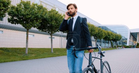 Téléchargez les photos : Portrait de l'homme barbu sérieux et confiant de bonne humeur dans des vêtements élégants qui descendent avec son vélo sur la rue pavée et parler sur smartphone - en image libre de droit
