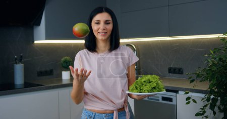 Téléchargez les photos : Attrayant sourire jeune brune heureuse debout dans la cuisine moderne et regardant caméra tenant dans les mains mangue et feuilles de laitue, concept de nutrition saine - en image libre de droit