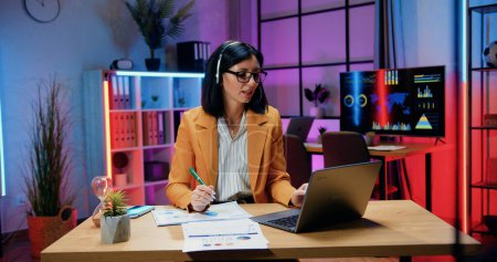 Téléchargez les photos : Concept d'entreprise où attrayant positif femme d'affaires qualifiée réussie dans casque et lunettes assis devant l'ordinateur pendant le chat en ligne avec des collègues - en image libre de droit