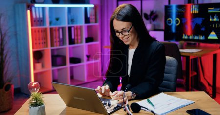Téléchargez les photos : Belle brune expérimentée et souriante en tenue élégante assise sur son lieu de travail au bureau du soir et tapant sur un ordinateur portable, concept d'entreprise - en image libre de droit