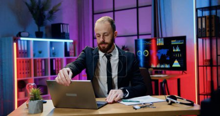 Téléchargez les photos : Beau concentré industrieux responsable habile homme d'affaires barbu assis sur son lieu de travail dans le bureau du soir et travaillant sur ordinateur - en image libre de droit