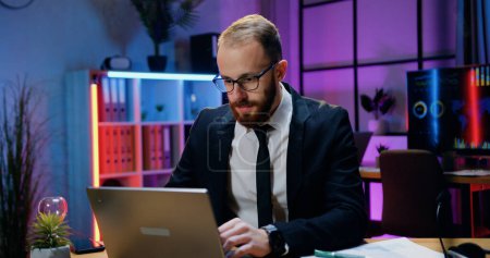 Téléchargez les photos : Confiance confiant travailleur qualifié homme d'affaires barbu en tenue formelle et dans des lunettes assis sur son lieu de travail et travaillant sur ordinateur à la fin du temps au bureau - en image libre de droit