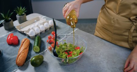 Téléchargez les photos : Femme au foyer méconnaissable ajoutant de l'huile d'olive dans une salade de légumes dans un bol en verre lors de sa préparation dans une cuisine contemporaine, concept de préparation maison - en image libre de droit