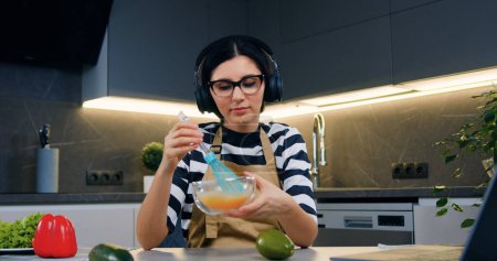 Photo for Lovely smiling happy young woman in earphones enjoying music while whipping eggs in bowl using whisk at the table in contemporary cuisine,front view - Royalty Free Image