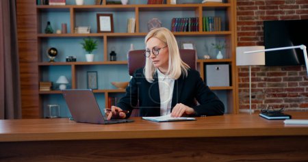 Téléchargez les photos : Attrayant concentré femme d'affaires blonde travaillant avec des informations sur les rapports d'ordinateur portable et d'affaires dans le bureau moderne - en image libre de droit