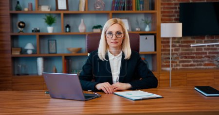 Téléchargez les photos : Portrait d'une belle femme d'affaires blonde adulte concentrée en tenue formelle assise devant la caméra dans un bureau moderne - en image libre de droit