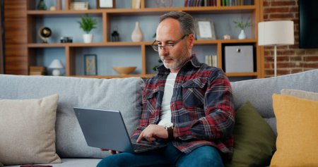 Photo for Good-looking skilled relaxed adult bearded man in glasses resting on comfortable sofa in sitting -room and surfing information on his computer - Royalty Free Image