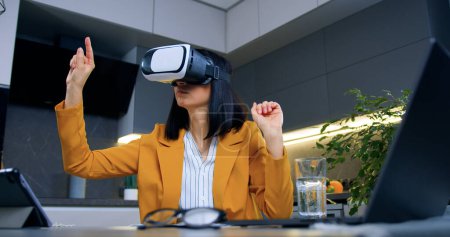 Téléchargez les photos : Femme aux cheveux bruns adulte entreprenante, confiante et confiante, en veste orange, assise à la table de la cuisine et travaillant à l'aide de lunettes spéciales 3D, au ralenti - en image libre de droit