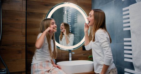 Téléchargez les photos : Les jeunes caucasiennes et les sœurs enfants se brossant les dents en se regardant en souriant. Smilling femme et enfant prenant soin de la santé dentaire le matin dans la salle de bain. - en image libre de droit