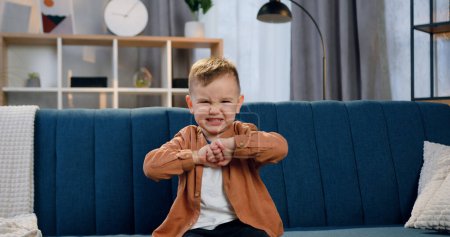 Téléchargez les photos : Adorable petit garçon drôle assis sur le canapé dans un appartement confortable et représentant la colère sur son visage en regardant dans la caméra - en image libre de droit