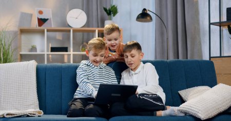 Foto de Primer plano de lindo feliz sonriendo tres chicos que se sientan en el cómodo sofá y viendo interesante programa en el ordenador - Imagen libre de derechos