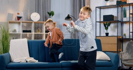 Foto de Preciosos chicos divertidos de 2 y 8 años divirtiéndose juntos en casa, el más pequeño saltando en el sofá, el mayor haciendo movimientos de baile - Imagen libre de derechos
