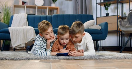 Portrait von attraktiven ruhigen 2-jährigen, 8-jährigen und 10-jährigen Jungen, die auf dem Boden liegen und die Überarbeitung des interessanten Programms auf dem Handy genießen