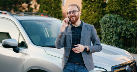Téléchargez les photos : Homme d'affaires souriant parlant sur son téléphone portable répondant à un appel d'affaires quand il se penche sur sa voiture qui est garée par la maison de la région. À l'extérieur - en image libre de droit