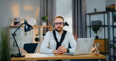 Photo for Handsome confident succssesful young bearded man in headset looking into camera sitting at his workplace in specially decorated home office - Royalty Free Image