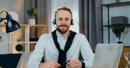 Photo for Close up of likable positive confident young bearded office worker in headphones which taking off his glasses while poses on camera in home office - Royalty Free Image