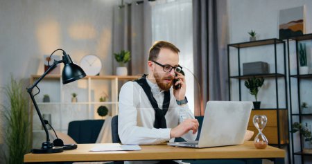 Photo for Likable experienced purposeful young bearded office worker in glasses talking on phone while working on computer in good-decorated home office - Royalty Free Image