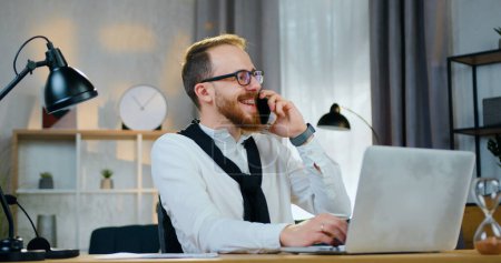 Photo for Handsome cheerful young bearded guy in glasses laughing from joke that hearing from his interlocutor on phone during working on laptop at home - Royalty Free Image