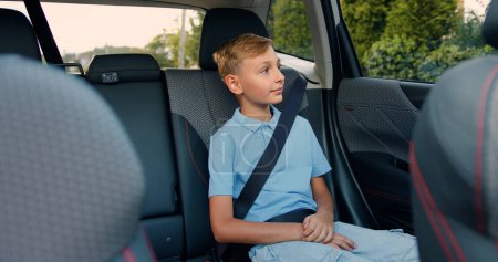 Téléchargez les photos : Petit garçon blond dans une chemise bleue ouvre la porte passager dans la voiture, s'assoit et attache sa ceinture de sécurité avant de commencer le voyage - en image libre de droit