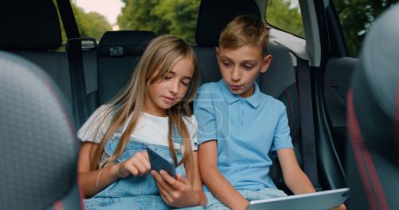 Téléchargez les photos : Enfants garçon et fille assis dans la voiture en utilisant une tablette numérique pour les jeux et les divertissements, célébrer le vin et donner haute cinq voyageant en voiture. Concept de vacances en famille. - en image libre de droit