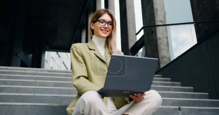 Photo for Good -looking young caucasian woman in glasses wearing a green jacket sitting with crossing legs on the stairs outside, typing on her laptop with satisfied face - Royalty Free Image