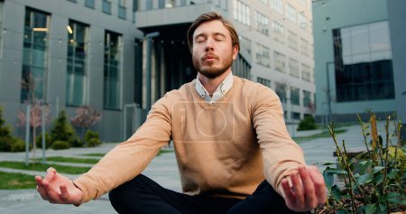 Photo for Handsome caucasian bearded man in glasses relaxing, sitting in the lotus position on the bench of the courtyard of office building or university. Healthy lifestyle, outdoors - Royalty Free Image