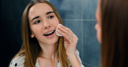 Close-up of young beautiful woman in pajamas with clean skin stands in bathroom in front of the mirror and removes makeup from her face with a cotton pad. Face portrait, facial skin care concept