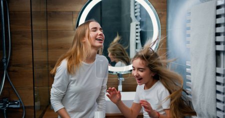 Foto de Dos chicas caucásicas divertidas con hermoso pelo largo bailando y cantando con secador de pelo en el baño moderno en casa. - Imagen libre de derechos