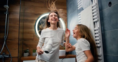 Foto de Mujer joven divertida y niña de 10 años con el pelo largo en pijama secando su pelo voluminoso por secador de pelo mientras bailan riendo y mirándose en el baño moderno en casa - Imagen libre de derechos