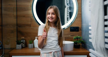 Téléchargez les photos : Portrait de sourire belle petite fille en pyjama avec les cheveux longs montrant pouce vers le haut, regardant la caméra tout en se tenant à l'arrière du miroir à la salle de bain dans la maison - en image libre de droit