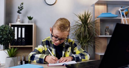 Photo for Front view of good-looking careful 10-aged modern boy in glasses which writing down the notes into notebook from computer - Royalty Free Image